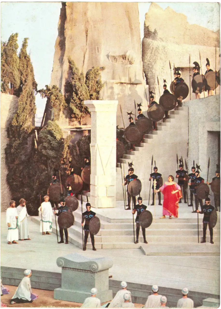 Fotografia a tutta pagina che ritrae una maestosa messa in scena teatrale dell'Antigone presso il Teatro Greco di Siracusa. Si vedono numerosi figuranti in armatura greca con lance e scudi disposti sulle scalinate di una scenografia imponente, con un attore in tunica rossa al centro e alte pareti di roccia sullo sfondo.